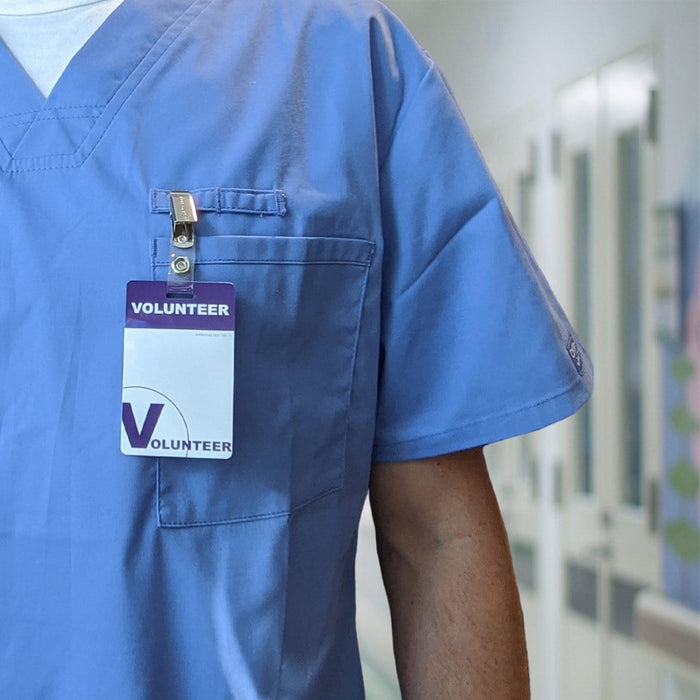 A person wearing blue scrubs with a 5 Pack - Purple Heavy Duty Plastic Volunteer Badges with Clothing Friendly Clip clipped to the front pocket stands in a hallway. Sold by Specialist ID.