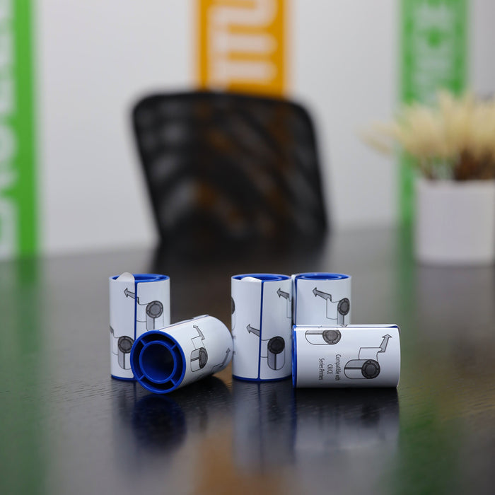 Six small, cylindrical containers with blue rims and illustrated labels—specifically 569946-002 Entrust (Datacard) Adhesive Cleaning Sleeves (5-Pack) for printhead protection—are neatly arranged on a dark table; a blurred chair and colorful wall panels are in the background.