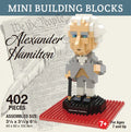 The Alexander Hamilton Mini Blocks set comes in a lightweight, glossy, and colorful cardboard box. The exterior features printed graphics, including a rendering of the assembled figure posed on a black base with a red platform beneath it. The product name appears in bold black script over a soft gray background depicting the U.S. Constitution and a white quill pen, tying the design to Hamilton’s legacy.