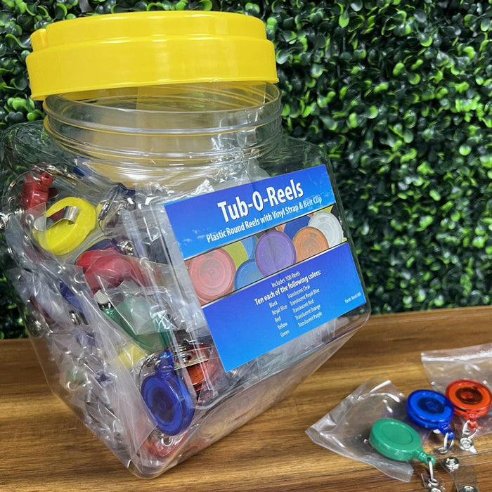 A plastic container labeled "Assorted Badge Reels TUB-O-REELS" is filled with various colored plastic round badge reels, set on a wooden surface with a green, leafy background. A few assorted badge reels are displayed outside the container, showcasing the variety of colors available.