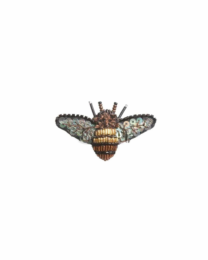 A small bee-shaped brooch with a rounded body and outstretched wings. The surface blends soft fabric with raised stitching, beads, and sequins, creating a gently textured feel with a subtle shimmer in shades of brown, blue, and black.The body is striped and slightly raised, with delicate wings extending outward. A smooth metal pin is attached to the back for fastening, and each handmade piece features slight variations in detail and finish.