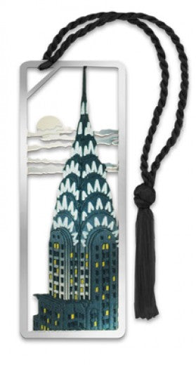 The image shows a silver-tone metal bookmark featuring a photo-etched Art Deco illustration of New York City’s Chrysler Building in green-blue, with yellow-lit windows. Above the skyscraper, stylized horizontal clouds stretch across a waning sun, and a small black tassel is attached at the top for decoration.
