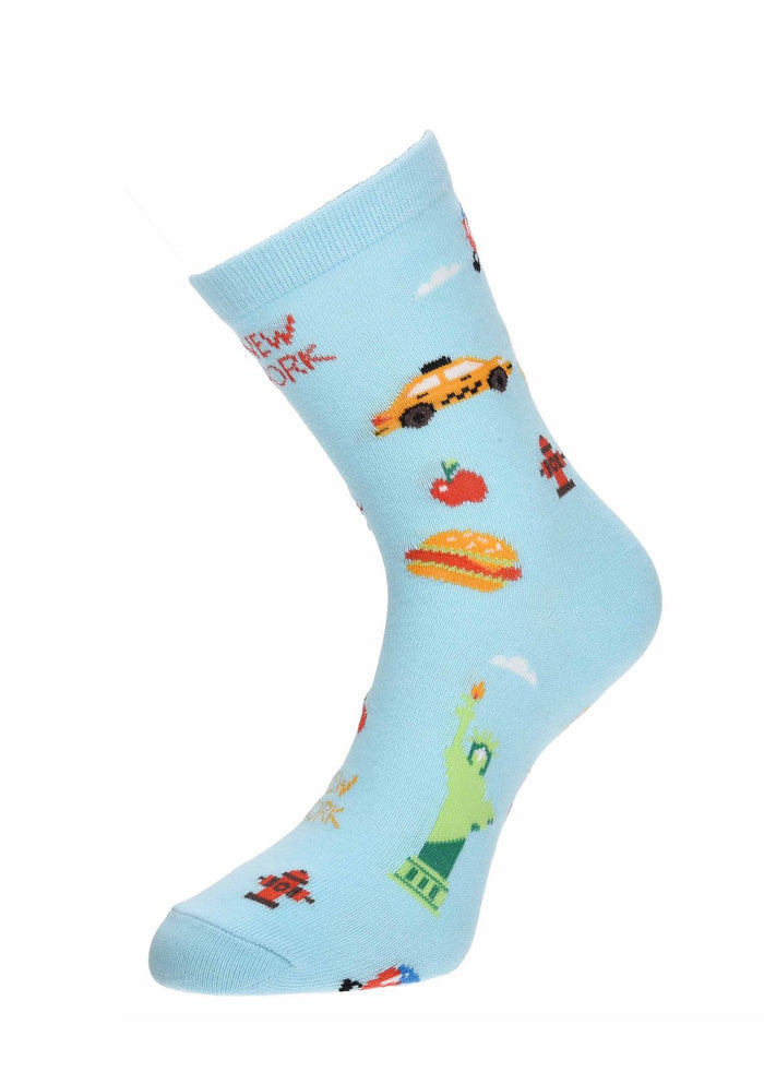 The image shows the light blue NYC Cartoon Icon Sock displayed on a mannequin foot. The sock is decorated with playful, colorful illustrations of New York City favorites, including the Statue of Liberty, yellow taxis, hot dogs, fire hydrants, and other iconic city symbols, as shown in the previous image.