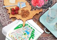 The image shows the Central Park Dish Towel displayed on a table beside drinks on a tray and a USA-shaped wooden cutting board topped with nuts and strawberries. The towel features a colorful illustrated map of Central Park, highlighting its landmarks, lakes, bridges, pathways, and attractions in a vibrant, hand-drawn style.