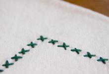 The image shows a corner of the dish towel, highlighting a border of hand-embroidered green X-stitches along the edge.