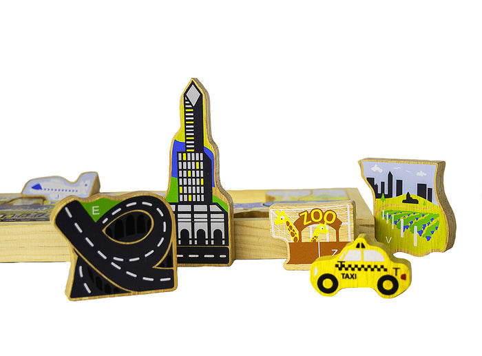 The image shows wooden cutout shapes of a skyscraper, a checkered taxi, a city skyline silhouette, and a black roadway with white lane markings. The pieces are painted in bright blues, greens, and yellows, with natural wood grain visible underneath. A smooth, shallow wooden tray appears in the background, holding the pieces neatly in place.