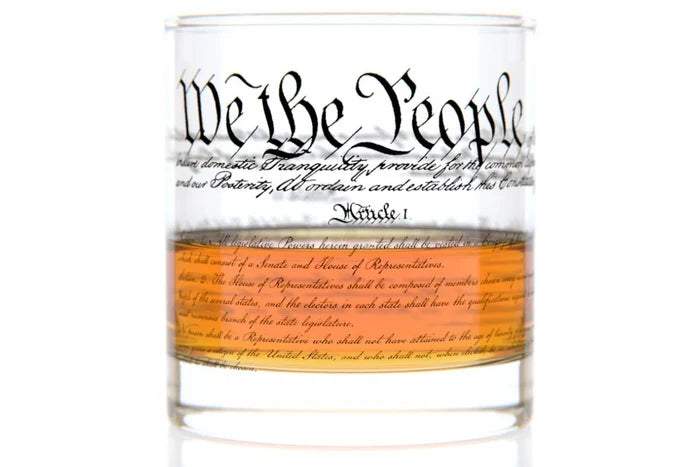 This 11 oz rocks glass is made of clear, heavy glass with a weighted base and a smooth, chip‑resistant rim. Near the top, “We the People” appears in period‑style cursive, while the exterior is wrapped with printed text of the U.S. Constitution. A clear orange band with additional cursive text encircles the lower half of the glass.