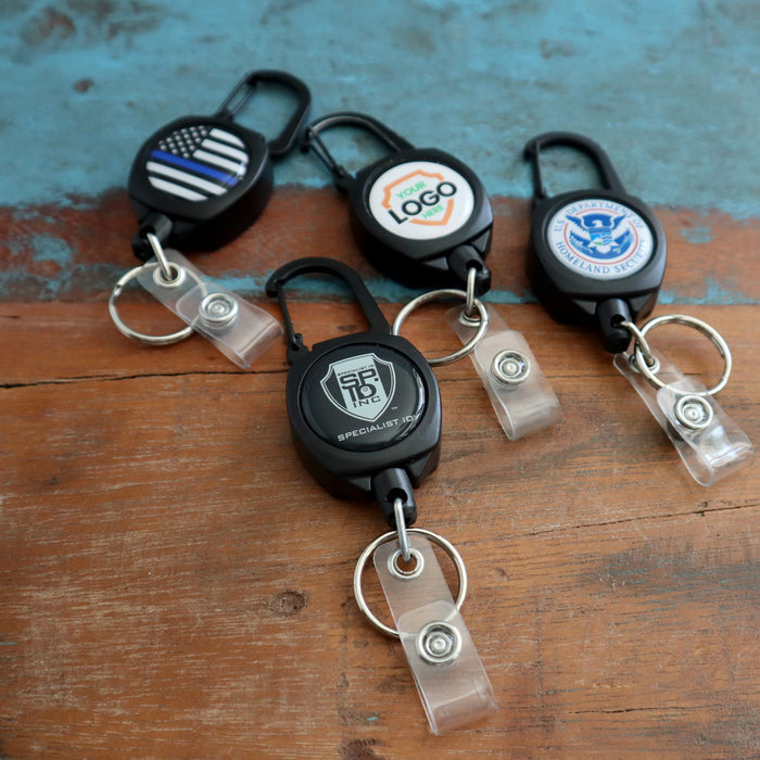 Four **Custom Compact Key-Bak Retractable Carabiner ID Badge Reels with Key Rings and Your Logo** are displayed on a wooden surface. Each Key-Bak holder features a different design or logo on its front, complete with durable retractable Kevlar cords.