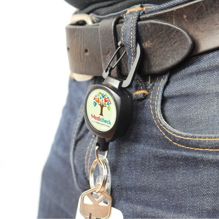 A black belt with a Custom Compact Key-Bak Retractable Carabiner ID Badge Reel with Key Ring and Your Logo clipped to it, displaying the MediCheck logo with a tree graphic. The retractable Kevlar cord holder has keys attached at the end, which are hanging over blue jeans.