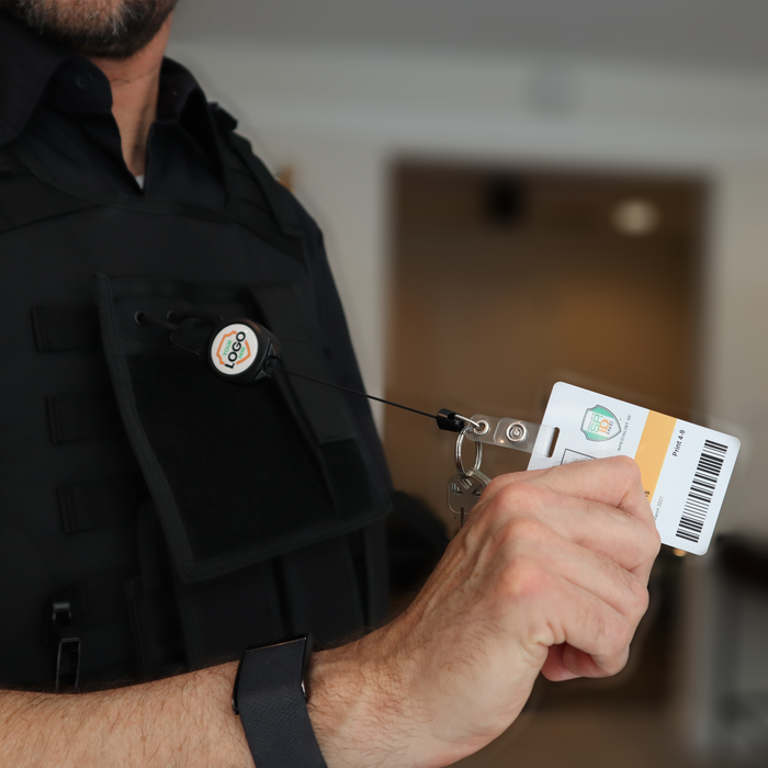 A person in a black uniform showcases an ID card connected to the Custom Compact Key-Bak Retractable Carabiner ID Badge Reel with Key Ring and Your Logo, enhancing durability. Sold by Specialist ID.