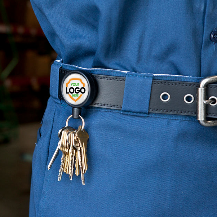 A set of keys attached to a Customizable Key-Bak Mid Size Key Ring Badge Reel with Belt Clip (#6) - Add Your Logo on the belt of a person wearing a blue uniform. The badge holder, which can be customized to promote brand awareness, has a placeholder for a logo.