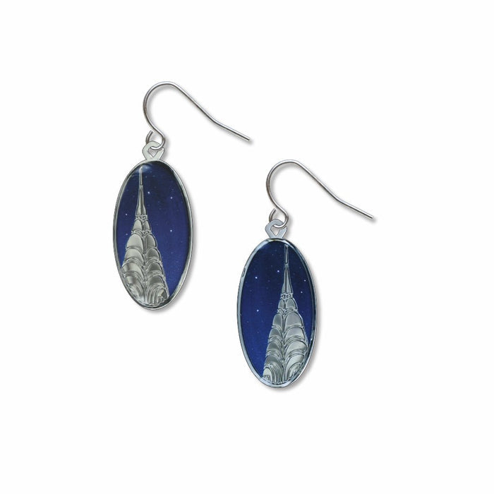 A pair of oval dangle earrings featuring a colorful Art Deco illustration of New York City’s Chrysler Building. The building’s illuminated silver-toned spire and signature tiered arches stand out against a deep lapis-blue night sky dotted with stars. The photo-etched giclée artwork is set within thin polished silver-plated brass frames and hangs from hypoallergenic ear wires. The earrings measure approximately 1.5 inches long.