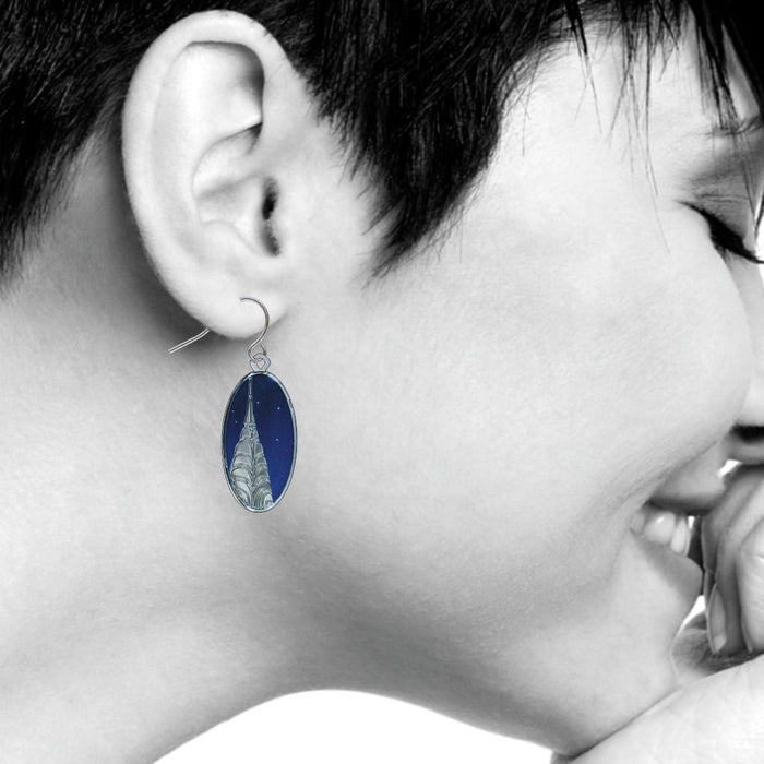 A black-and-white photograph shows a model in profile wearing one of the Chrysler Building at Night dangle earrings. The oval earring, shown in color against the monochrome image, hangs below the ear and features an Art Deco illustration of the Chrysler Building’s silver-toned spire and tiered arches set against a starry lapis-blue night sky.