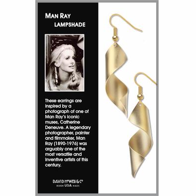 Lampshade Man Ray Spiral Earrings in matte gold displayed in packaging with an information card featuring a photograph of French actress Catherine Deneuve, Man Ray’s iconic muse. The dangle earrings feature open spiral cone shapes inspired by a Man Ray photograph and hang from gold wire hooks.