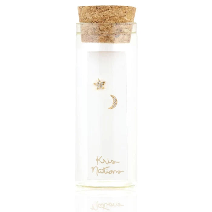 A small clear glass vial with a cork stopper sits against a white background. Inside is a pair of petite gold stud earrings—one shaped like a crescent moon and the other like a star. Both are covered in tiny sparkling clear crystals that catch the light.