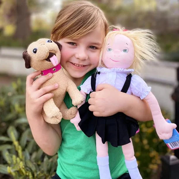The image shows a smiling young girl holding the fully dressed 18‑inch Eloise doll, along with a plush toy dog. They are posed outdoors against a leafy green background, creating a warm, cheerful scene that highlights the doll as a cuddly companion.