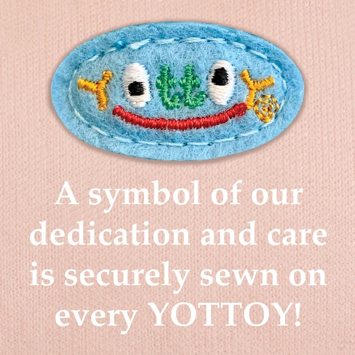 The image shows an embroidered blue YOTTOY logo in multicolored stitching with a small red smile, sewn onto pink knit fabric. Below the emblem, bold white text reads: “A symbol of our dedication and care is securely sewn on every YOTTOY!”