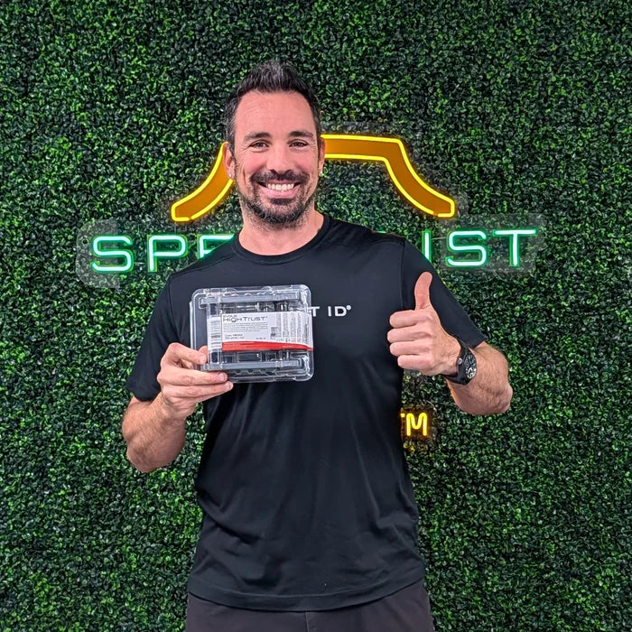 A man in a black t-shirt stands before a green, plant-covered wall with neon signage, holding a clear box—highlighting the vibrant full-color prints produced by the Evolis R5F208A100 YMCKO Ribbon for Primacy 2 card printers—and gives a thumbs-up.