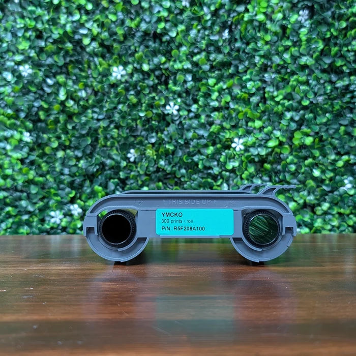 An Evolis R5F208A100 YMCKO Color Ribbon (300 prints, P/N: R5F208A100) sits on wood with a leafy backdrop—perfect for vibrant, photo-quality Primacy 2 ID card printing with overlay protection.