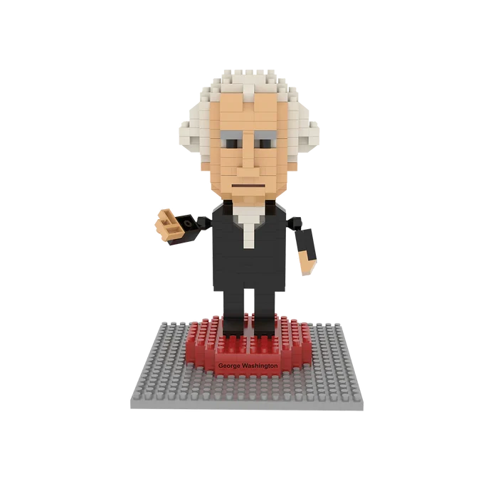 A toy figure of George Washington stands upright on a small base labeled with his name, set against a plain white background. The figure is about 4.5 inches tall and made from tiny interlocking blocks, wearing a long, black 18th-century coat with simple, calm facial features.