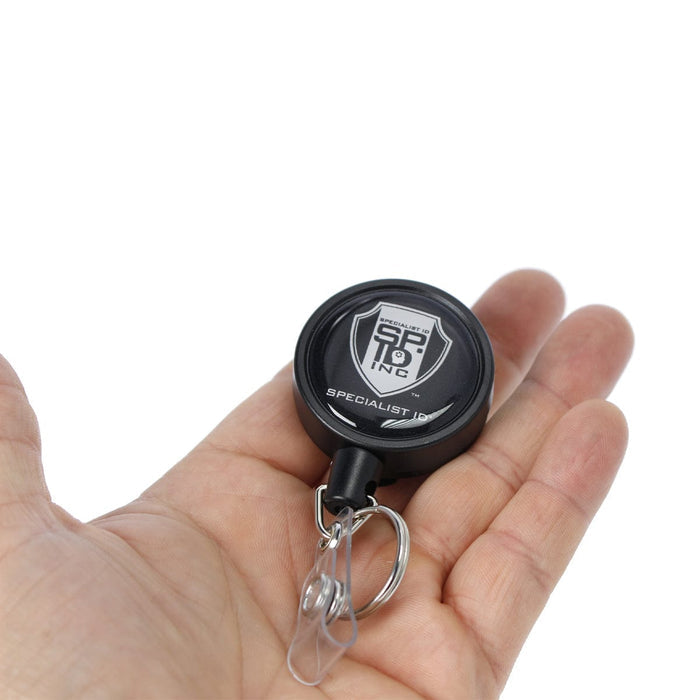 A hand holding a keychain with a Heavy Duty Badge Reel w/ Metal Swivel Belt Clip & Key Ring featuring a logo and the text "Specialist ID Inc.," complemented by a durable metal swivel belt clip for secure fastening.