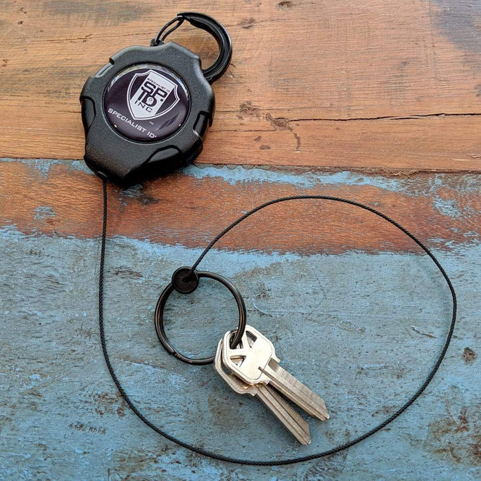 Heavy Duty Retractable Ratchit Keychain Tether Reel for Multiple Keys with Clip and Locking Feature with a logo, featuring two silver keys attached by a ring, placed on a wooden surface with blue and brown paint.