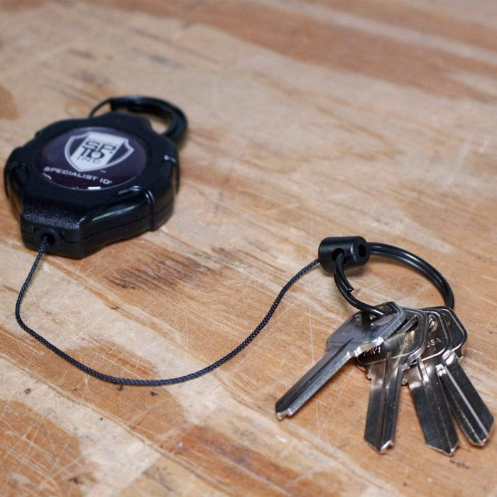 A set of keys attached to a Heavy Duty Retractable Ratchit Keychain Tether Reel for Multiple Keys with Clip and Locking Feature lies on a wooden surface. Sold by Specialist ID.