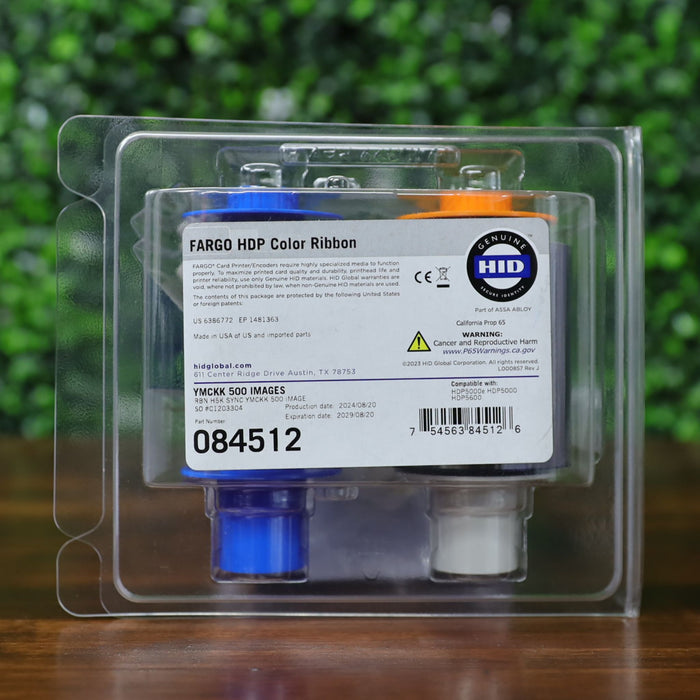 Close-up front view of the HID Fargo 084512 YMCKK printer ribbon in its clear plastic clamshell packaging, showing the full product label with barcode, part number, ribbon type, and manufacturer warnings. Label indicates 500 image yield, dual black panels, and retransfer compatibility with HDP5600 and HDP5000 series printers. Photographed and sold by Specialist ID, an authorized HID Fargo dealer.