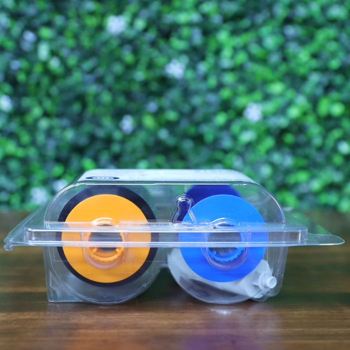 Top-down view of the Fargo 084512 YMCKK ribbon lying flat in its clamshell package, clearly showing the color-coded reels—orange for YMC panels and blue for resin black panels—positioned horizontally inside. Useful for identifying correct ribbon orientation prior to printer installation. Photographed and sold by Specialist ID, an authorized HID Fargo dealer.