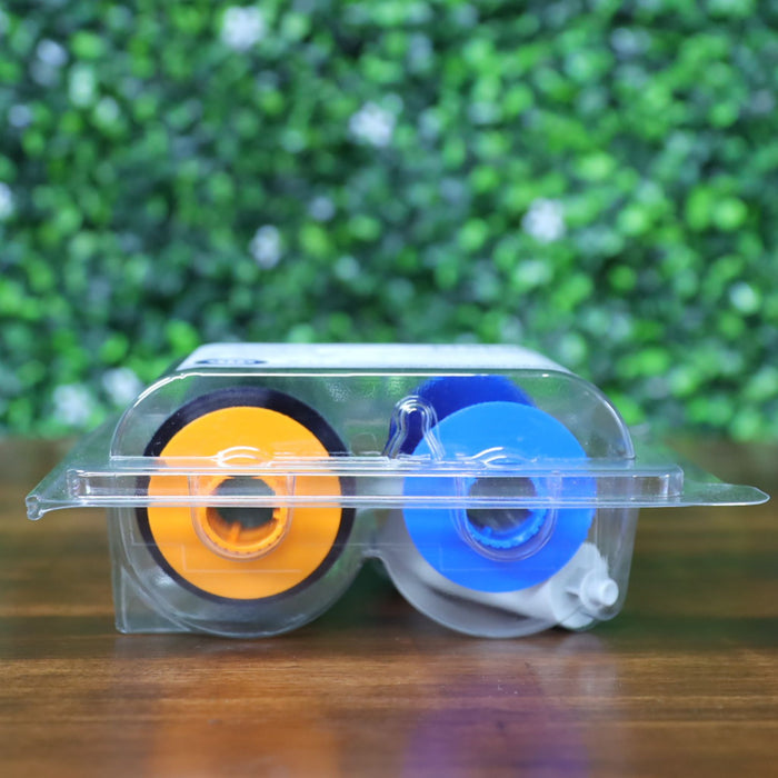 Top-down view of the HID Fargo 84514 YMCFK ribbon lying flat inside its factory-sealed clamshell. The orange and blue spool hubs are visible through the clear plastic, with the white cleaning roller positioned beside them. Photographed and sold by Specialist ID, an authorized HID Fargo dealer.