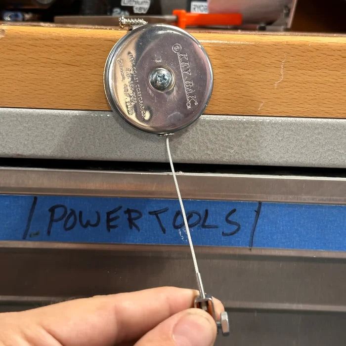 A hand holding a retractable reel labeled "Key-Bak #481LS-HD Mountable Retractable Reel with Steel Cable and L Bracket + Hex Nut Yoke" against a drawer marked "Power Tools" on blue tape.