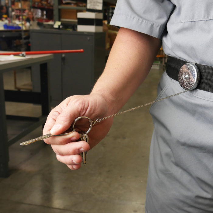 A person wearing a gray uniform holds keys attached to a Key-Bak #5 Self Retracting Key Badge Reel clipped onto their belt in a workspace. Sold by Specialist ID.