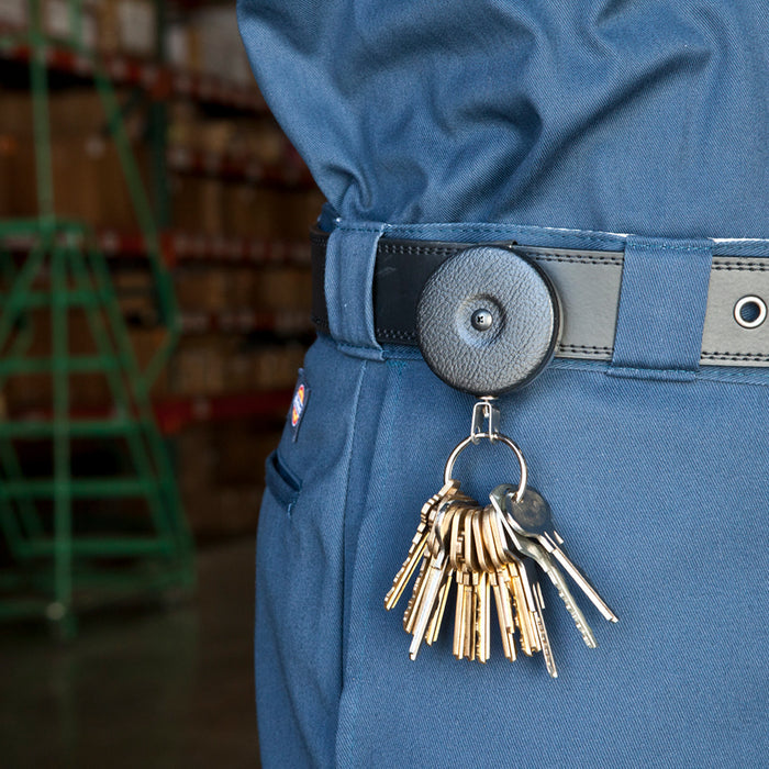 A close-up of a person wearing navy blue pants with a black belt holding a Key-Bak #5B Self Retracting Heavy Duty Key/Badge Reel with several keys attached, standing in a warehouse environment.