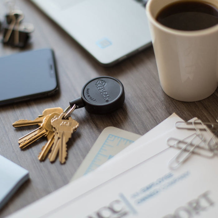 A set of keys on a key ring, a Key-Bak Mid Size Key Ring Badge Reel with Belt Clip (#6) attached to a belt clip, a smartphone, a laptop, a cup of coffee, and papers with paper clips are all meticulously arranged on the wooden surface.