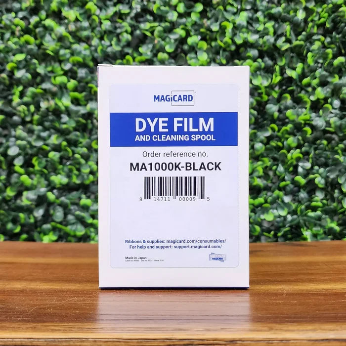 A Magicard MA1000K-Black Monochrome K Ribbon for ID card printers (1000 prints) sits on a wooden surface with a leafy green background.