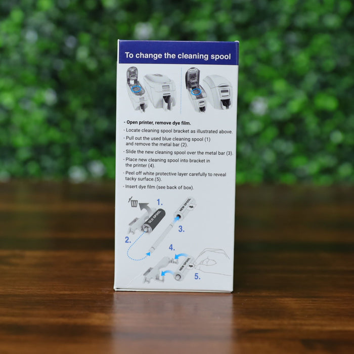 Side of Magicard MA1000K-Blue ribbon retail box showing illustrated instructions for changing the cleaning spool, displayed on a wood surface with green leafy background. Photographed and sold by Specialist ID, an authorized Magicard dealer.