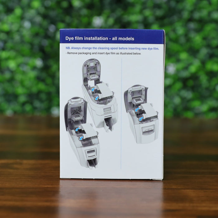 Back of Magicard MA1000K-Blue ribbon retail box showing illustrated dye film installation instructions for all models, displayed on a wood surface with green leafy background. Photographed and sold by Specialist ID, an authorized Magicard dealer.