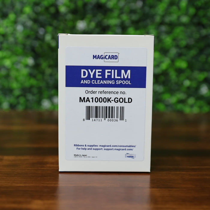 A white box labeled "Magicard MA1000K GOLD Gold Ribbon – 1000 Prints," order reference MA1000K-GOLD, is on a wooden surface with green leaves in the background—product description text is absent, replaced by CSS code.  Photographed and sold by Specialist ID, an authorized Magicard dealer.