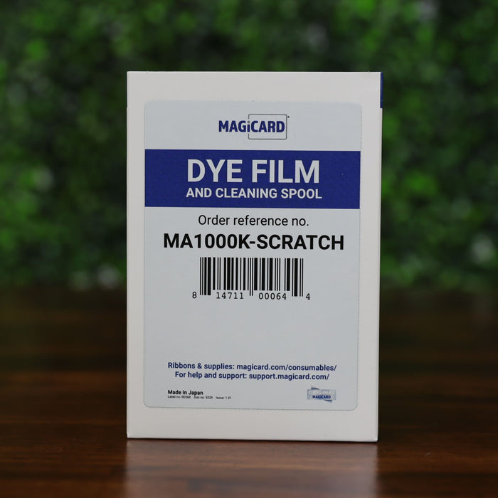 Magicard MA1000K-Scratch scratch-off ribbon box with dye film and cleaning spool label, displayed on a wood surface with green leafy background, sold by Specialist ID, an authorized Magicard dealer