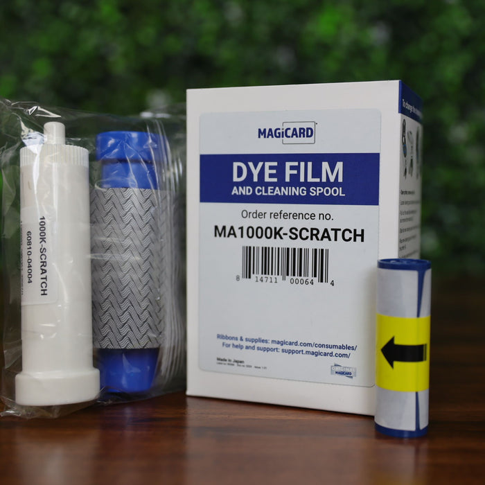 Magicard MA1000K-Scratch scratch-off ribbon kit including ribbon cartridge with wavy gray pattern, cleaning spool, and retail box, displayed on a wood surface with green leafy background, photographed and sold by Specialist ID, an authorized Magicard dealer.