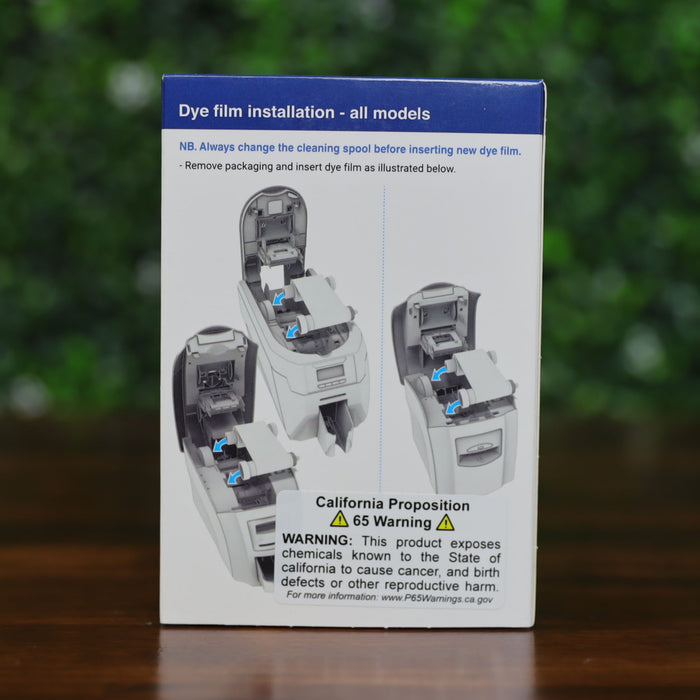 Back of Magicard MA1000K-Scratch scratch-off ribbon box showing step-by-step illustrated instructions for replacing the cleaning spool, with text and diagrams, displayed on a wood surface with green leafy background, photographed and sold by Specialist ID, an authorized Magicard dealer.
