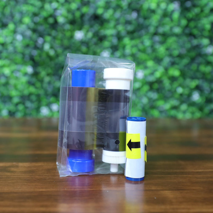 Magicard MB300YMCKO Color Ribbon (300 prints) ribbon cartridge and cleaning spool shown sealed in clear plastic with a separate blue arrow cleaning roller, placed on a wooden surface with a blurred green foliage background. Photographed and sold by Specialist ID, an authorized Magicard dealer.