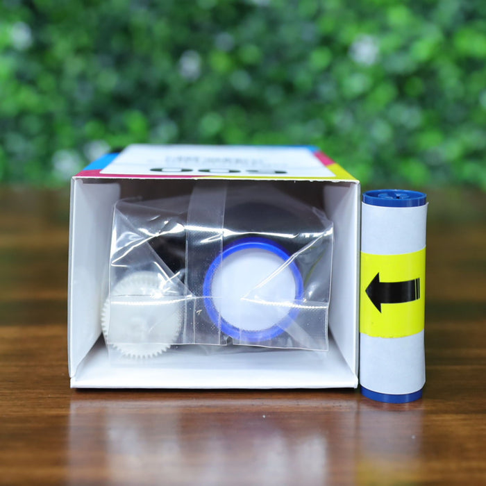 Magicard MB300YMCKO Color Ribbon (300 prints) box lying down on its side, open to show the blue and white ribbon inside sealed in clear plastic, with a separate blue arrow cleaning roller beside it, on a wooden surface with a blurred green leafy background. Photographed and sold by Specialist ID, an authorized Magicard dealer.