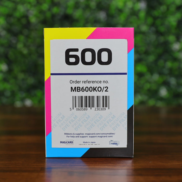 A Magicard MB600KO/2 ribbon box labeled 600 with a barcode on a wood table in front of a green leafy background, photographed and sold by Specialist ID.