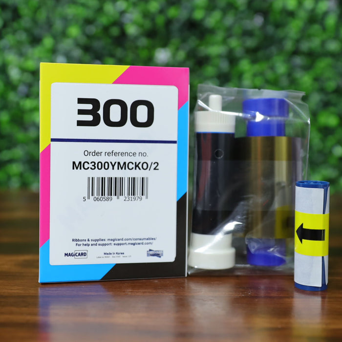 Magicard MC300YMCKO color ribbon and packaging for 300 ID card prints, shown with cleaning spool and ribbon roll on a wood surface with green background, photographed and sold by Specialist ID, an authorized Magicard dealer.