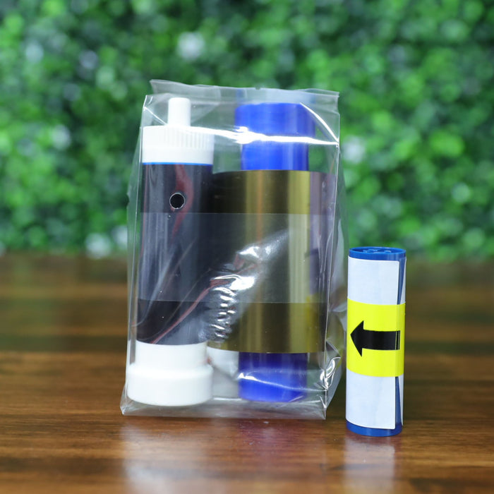 A sealed plastic bag containing a Magicard MC300YMCKO color ribbon and white cleaning spool sits on a wood surface, with a separate ribbon roll marked with a yellow arrow beside it, against a green leafy background. Photographed and sold by Specialist ID, an authorized Magicard dealer.