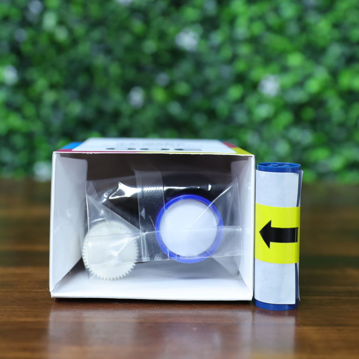 A kit box containing a blue-capped ribbon, white cleaning spool, and a separate rolled ribbon marked with a yellow arrow for the Magicard MC300YMCKO color ribbon (300 prints) sits on a wooden surface with a green leafy background. Ideal for Magicard 300 printer users needing ribbon replacement. Photographed and sold by Specialist ID, an authorized Magicard dealer.