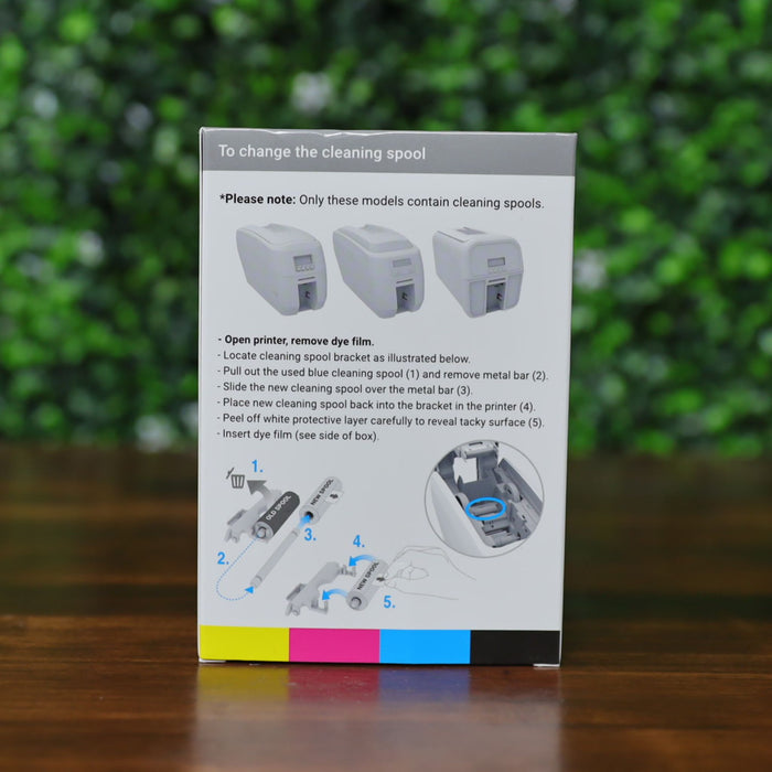Box containing the Magicard MC300YMCKO YMCKO Color Ribbon (300 Prints) with step-by-step illustrated installation instructions, displayed on a wooden surfBack of Magicard MC300YMCKO color ribbon box (300 prints) showing step-by-step illustrated cleaning spool and dye film installation instructions, with diagrams and text on a white panel, displayed on a wood surface with a blurred green leafy background. Photographed and sold by Specialist ID, an authorized Magicard deace against a blurred green background.