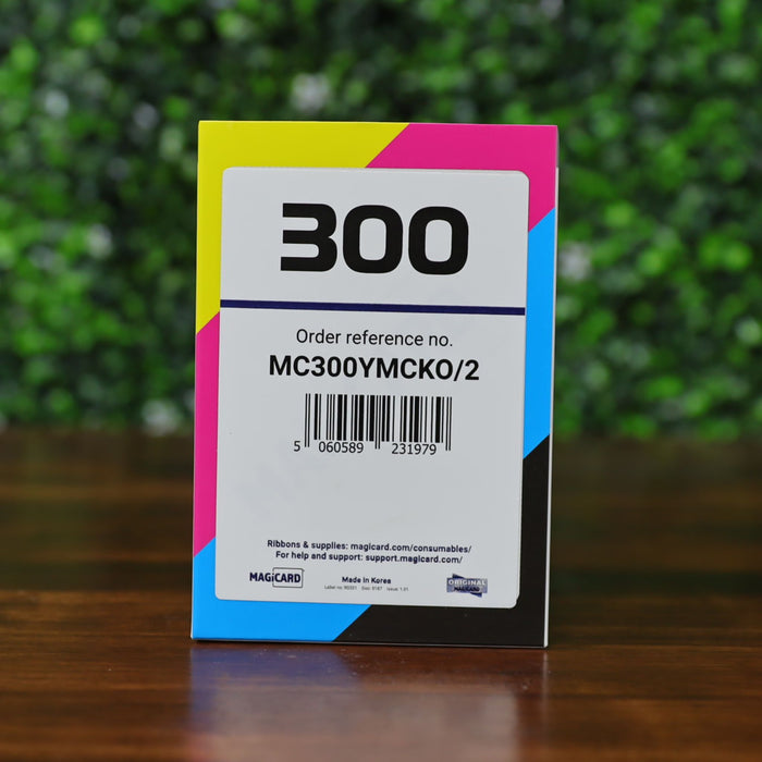 Front of Magicard MC300YMCKO color ribbon box for 300 prints, order reference MC300YMCKO/2, designed for the Magicard 300 ID card printer, displayed on a wood surface with a blurred green leafy background. Photographed and sold by Specialist ID, an authorized Magicard dealer.
