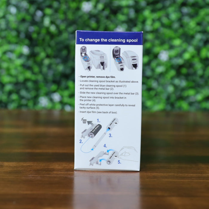 Box with step-by-step illustrated instructions for changing a Magicard PR1000GL Monochrome Gold Ribbon (1000 prints), compatible with PriceCardPro 100 and Flex+ ID card printers, set against a blurred green and brown background. Photographed and sold by Specialist ID, an authorized Magicard dealer.

—